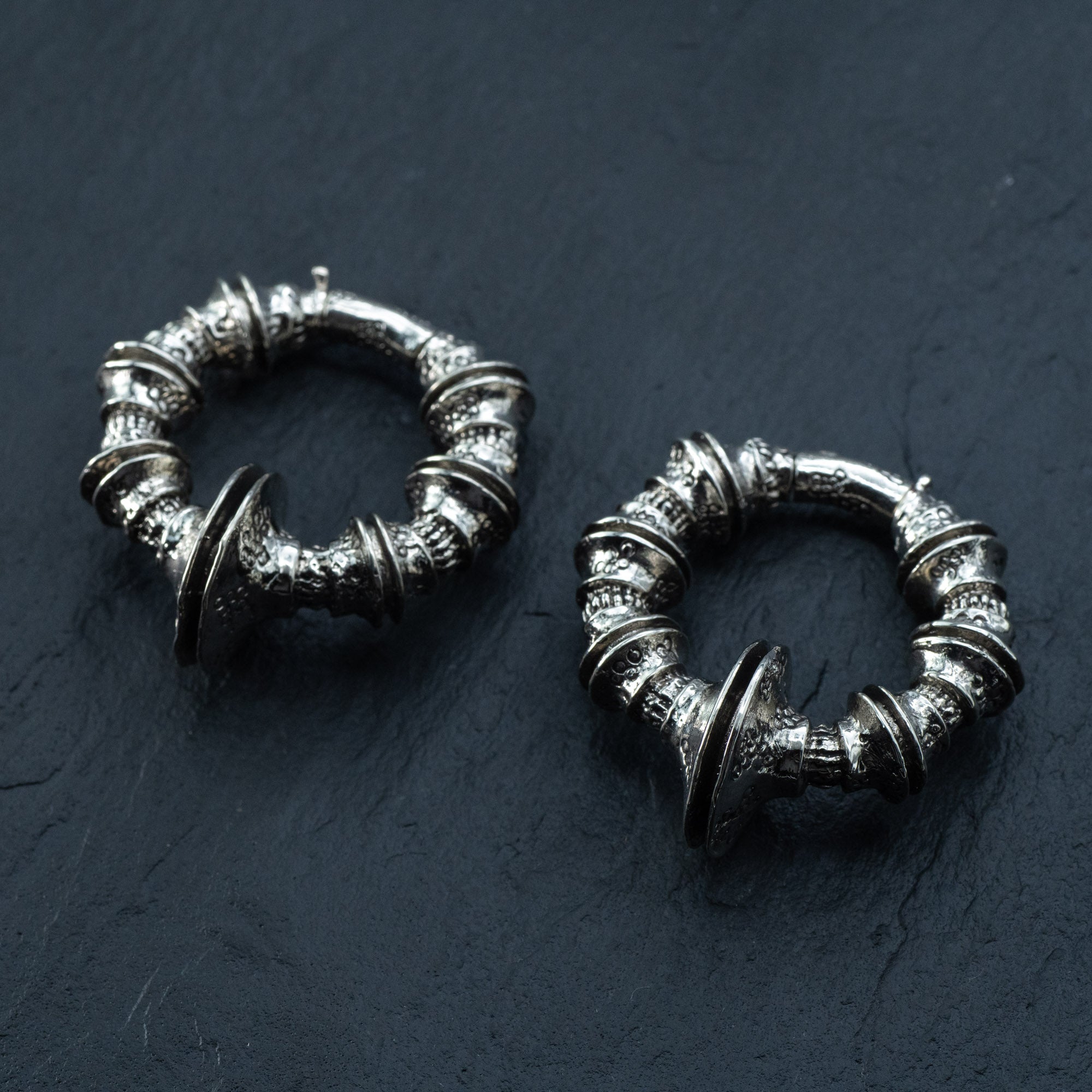 The TRAKARA Heavy Tribal Cyborg Clicker Ear Weights in Silver from Kaya Movement, designed intricately with circular shapes and detailed embellishments, rest on a dark, textured surface. These 2 gauge earrings showcase a vintage, antique aesthetic with ornate patterns and a slightly oxidized finish, reminiscent of tribal cyborg earrings.